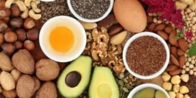 Vista superior de alimentos saudáveis como abacate, nozes, sementes, ovos e castanhas dispostos em tigelas.