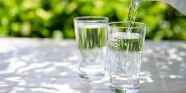 Água sendo servida em copo de vidro sobre mesa branca, com fundo verde desfocado de plantas ao sol.