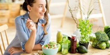 Mulher sorrindo à mesa com salada e sucos naturais, simbolizando escolhas saudáveis e consciência alimentar.