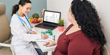 Em um consultório, uma médica está atendendo uma mulher obesa. Ambas estão sentadas em volta de uma mesa, conversando.