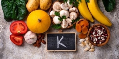 Alguns alimentos ricos em potássio, junto a um quadro negro com a letra K desenhada, representando potássio para que serve.