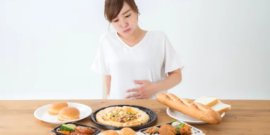 Mulher sentada à mesa com vários pratos de comida, segurando a barriga, representando saciedade.
