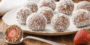 Sobremesas fit Docinhos fit de morango cobertos com coco ralado em prato branco sobre mesa rústica de madeira.