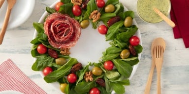 Guirlanda de salada com folhas verdes, tomatinhos, azeitonas, nozes, tâmaras e flor de salame ao centro.