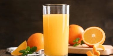 Copo de suco de laranja natural sobre mesa rústica, com laranjas inteiras e cortadas ao fundo.