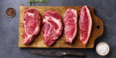 Cortes de carne bovina crua dispostos sobre tábua de madeira, com faca, sal grosso e alecrim, representando carnes para bife.