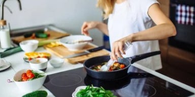 Mulher cozinhando em frigideira sobre cooktop, com ingredientes frescos organizados na bancada da cozinha.