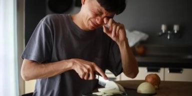 Pessoa cortando cebola sobre tábua de madeira na cozinha, demonstrando que está chorando.