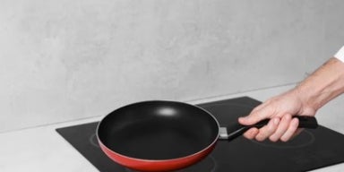 Mão segurando frigideira antiaderente sobre cooktop de indução, pronta para fazer a cura da panela.