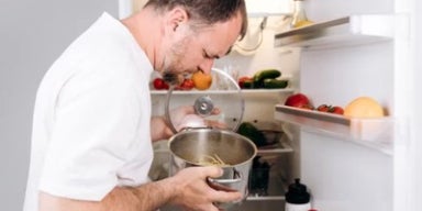 Homem observa panela com comida da geladeira aberta, observando ingredientes e recipientes nas prateleiras internas.