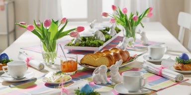 Decoração de Páscoa: Mesa decorada com pratos, talheres, pães, vaso com flores e xícaras