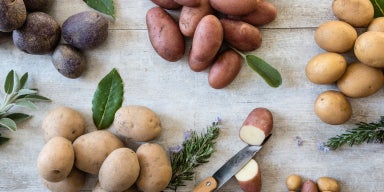 Seleção de batatas vermelhas, amarelas e roxas acompanhadas de ervas aromáticas sobre superfície de madeira rústica.