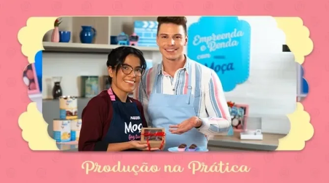 Fotografia com tons rosa, azul e amarelo mostra duas pessoas sorrindo enquanto mostram uma caixinha com brigadeiros.