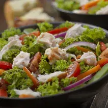Fotografia em tons de verde, em uma bancada de madeira de cor marrom. Ao centro, um prato redondo preto contendo a salada e ao fundo, um outro prato contendo salada. Ao lado, há um garfo de inox.