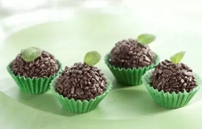 fotografia em tons de verde e marrom, contém 4 brigadeiros com granulados por cima dentro de forminhas verdes e todos tem uma folha de hortelã por cima para decorar fotografia em tons de verde e marrom, contém 4 brigadeiros com granulados por cima dentro de forminhas verdes e todos tem uma folha de hortelã por cima para decorar