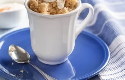 Fotografia em tons de branco e azul, com caneca branca ao centro contendo bolo e uma colher sobre prato azul, ao lado guardanapo em branco e azul, acima potinho com iogurte, tudo sobre bancada em tons de azul.