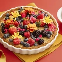 Foto da receita de Torta de Iogurte. Observa-se a torta decorada de frutas vermelhas e flores comestíveis sobre uma superfície vermelha. Ao lado direito, biscoitos inteiros decoram a foto.