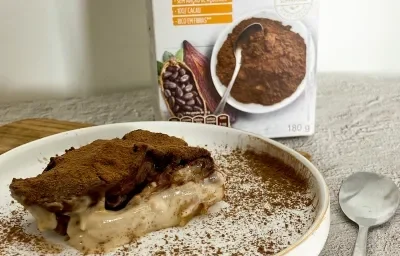 Foto da receita de Bombom de travessa plant-based. Observa-se um pedaço da torta em um prato branco polvilhado de cacau em pó e com com uvas decorando. Foto da receita de Bombom de travessa plant-based. Observa-se um pedaço da torta em um prato branco polvilhado de cacau em pó e com com uvas decorando.