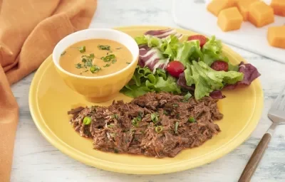 Foto da receita de Purê de Abóbora com Carne Desfiada. Observa-se um prato amarelo com a carne desfiada e a salada e, dentro de um potinho amarelo, o purê de abóbora finalizado com salsinha.