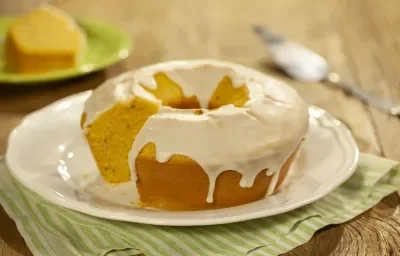 Fotografia em tons de marrom, verde e branco de uma bancada de madeira com um paninho verde, sobre ele um prato redondo branco com um bolo de fubá. Ao fundo uma espátula e um prato redondo verde com uma fatia de bolo.