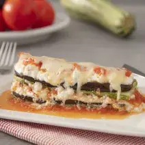 Foto da Receita de Lasanha de Ratatouille com Creme de Ricota. Observa-se um prato quadrado branco com uma fatia da lasanha. Atrás, de fundo, tomates e abobrinha decoram a foto.