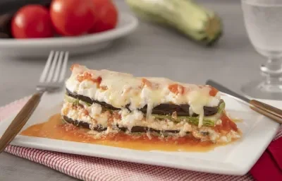 Foto da Receita de Lasanha de Ratatouille com Creme de Ricota. Observa-se um prato quadrado branco com uma fatia da lasanha. Atrás, de fundo, tomates e abobrinha decoram a foto. Foto da Receita de Lasanha de Ratatouille com Creme de Ricota. Observa-se um prato quadrado branco com uma fatia da lasanha. Atrás, de fundo, tomates e abobrinha decoram a foto.