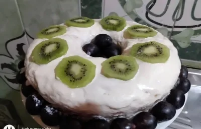 Foto da receita de bolo de cenoura confeitado servido com calda de chantilly, rodelas de kiwi cortadas por cima e uvas rosas para decorar em volta