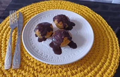 Fotografia em tons de amarelo em uma bancada de pedra, um porta prato amarelo de renda, um prato branco com bolinhos de cenoura com cobertura de chocolate por cima.