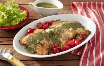 Foto da receita de frango caprese servida com tomates cereja em um prato largo e fundo de porcelana branca sobre uma mesa de madeira com um pano vermelho listrado a lado