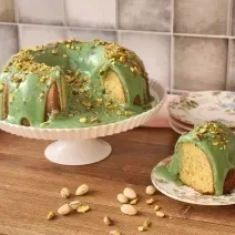 Fotografia em tons de verde em uma bancada de madeira, com um bolo vulcão de pistache ao centro. Ao lado, um prato com uma fatia do bolo e alguns pratos de cerâmica rosa.