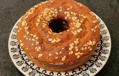 Foto da receita de Bolo integral. Observa-se um bolo assado com aveia em flocos por cima em um prato preto e branco decorado.