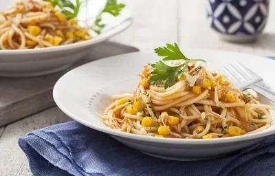 Fotografia em tons de branco e azul de uma bancada branca, com um pano azul dobrado e em cima dele um prato redondo e fundo branco com macarrão, 2 folhas de salsa e um garfo. Ao fundo, outro prato de macarrão e um copo azul e branco.