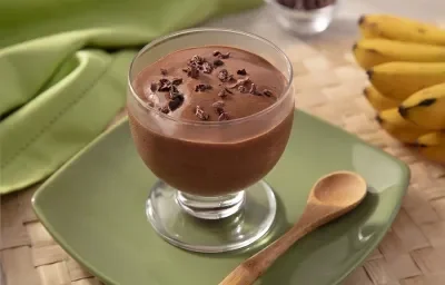 Fotografia em tons de marrom em uma bancada de madeira de cor amarela. Ao centro, um prato verde contendo uma taça com a mousse. Ao lado, há uma colher de madeira e ao fundo, um pano verde e um cacho de banana. Fotografia em tons de marrom em uma bancada de madeira de cor amarela. Ao centro, um prato verde contendo uma taça com a mousse. Ao lado, há uma colher de madeira e ao fundo, um pano verde e um cacho de banana.