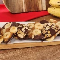 Fotografia em tons de vermelho em uma bancada de madeira com uma tábua e em cima barras de banana com chocolate e amêndoas . Ao fundo, um cacho de banana e um pano vermelho.