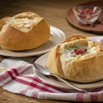 Foto de um pão italiano com sopa creme de queijo dentro e bacon decorando em cima da receita