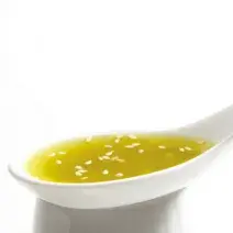 Fotografia em tons de amarelo em um fundo e mesa brancos com uma colher com o molho de mostarda com gergelim.