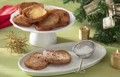 Foto da receita de rabanada sem glúten servida sobre uma base de porcelana branca com duas fatias separadas polvilhadas com açúcar de confeiteiro. Ao fundo há um paninho vermelho e decoração natalina Foto da receita de rabanada sem glúten servida sobre uma base de porcelana branca com duas fatias separadas polvilhadas com açúcar de confeiteiro. Ao fundo há um paninho vermelho e decoração natalina