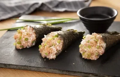 Foto da receita de Temaki de Salmão. Observa-se 3 temakis em cima de uma tábua de preta e, atrás, um potinho com molho shoyu e um ramo de cebolinha.