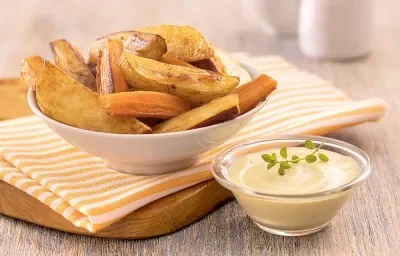 Foto em tons de amarelo da receita de legumes rústicos com molho rosé servida em um bowl de porcelana branco sobre um pano listrado laranja em cima de uma tábua de madeira. À frente há um recipiente de vidro com o molho rosé