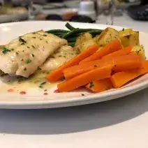Fotografia em tons de branco em uma bancada de madeira de cor branca. Ao centro, um prato contendo um filé de pescada com legumes ao lado.