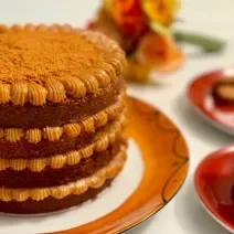 Fotografia em tons de caramelo com um prato de cor marrom ao centro. Em cima do prato existe um bolo com várias camadas de doce de leite e coberto com biscoito triturado