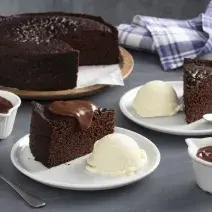 Fotografia em tons de marrom e azul de uma bancada cinza, ao centro dois pratos brancos com fatias de bolo de chocolate e ao lado uma bola de sorvete de creme. Ao fundo o bolo inteiro sem duas fatias.
