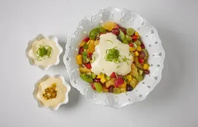 foto em tons de branco de uma bancada branca vista de cima, contém um recipiente redondo branco ao centro com pedaços de frutas e creme. Ao lado dois potinhos redondos brancos com creme.