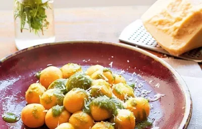 Fotografia em tons de vinho e laranja, ao centro prato vinho com bolinhas alaranjadas cobertas com molho verde e queijo ralado sobre guardanapo cinza, no entorno, um garfo, pedaço de queijo sobre ralador, vasinho com manjericão, tudo sobre bancada marrom.