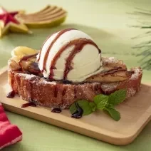 Foto da receita de rabanada vegana servida sobre com uma bola de sorvete por cima sober uma base retangular de madeira em cima de uma mesa com decoração de natal e um paninho vermelho ao lado