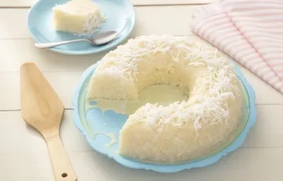 Fotografia em tons de azul e rosa de uma bancada de madeira branca, ao centro um prato azul com o cuscuz de tapioca coberto com coco ralado. Ao lado uma espátula de madeira em um pano com listras rosas e brancas.