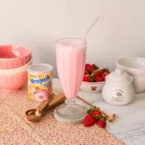 Fotografia de um milkshake rosa servido em uma copo alto de vidro. Ao fundo um cenário com morangos e bowls com tons rosas.