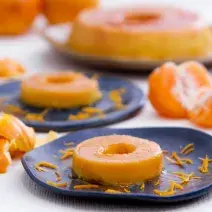 Imagem aproximada de um pudim feito com casca de mexerica, em tom laranja claro, sobre um prato azul escuro numa mesa com alguns gomos de mexerica