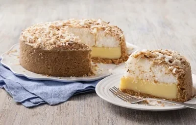 Fotografia em tons de marrom e azul de uma bancada de madeira com um paninho azul, sobre ele um prato branco com uma torta de coco. Ao lado um prato pequeno redondo com uma fatia da torta e um garfo.