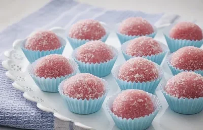 Fotografia em tons de azul e rosa de um paninho azul, sobre ele um prato redondo com decoração de florezinhas, sobre ele 12 brigadeiros rosa, com açúcar polvilhado, em forminhas azuis claras para doce.